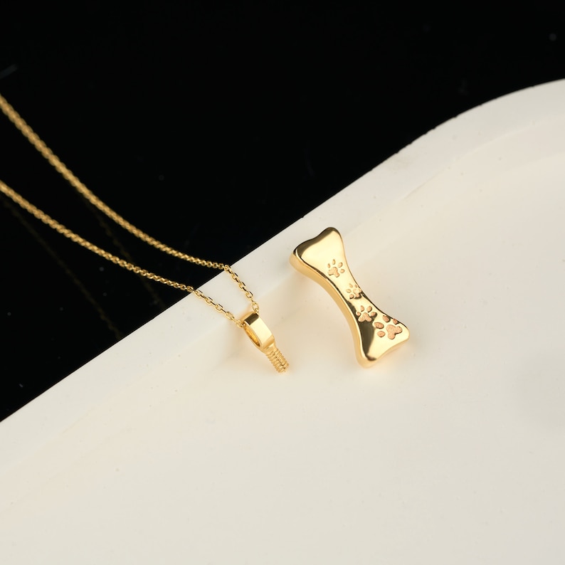 May include: A gold-colored necklace with a small pendant and a separate bone-shaped charm. The bone charm has paw prints. The necklace and charm are displayed on a white surface with a black background.