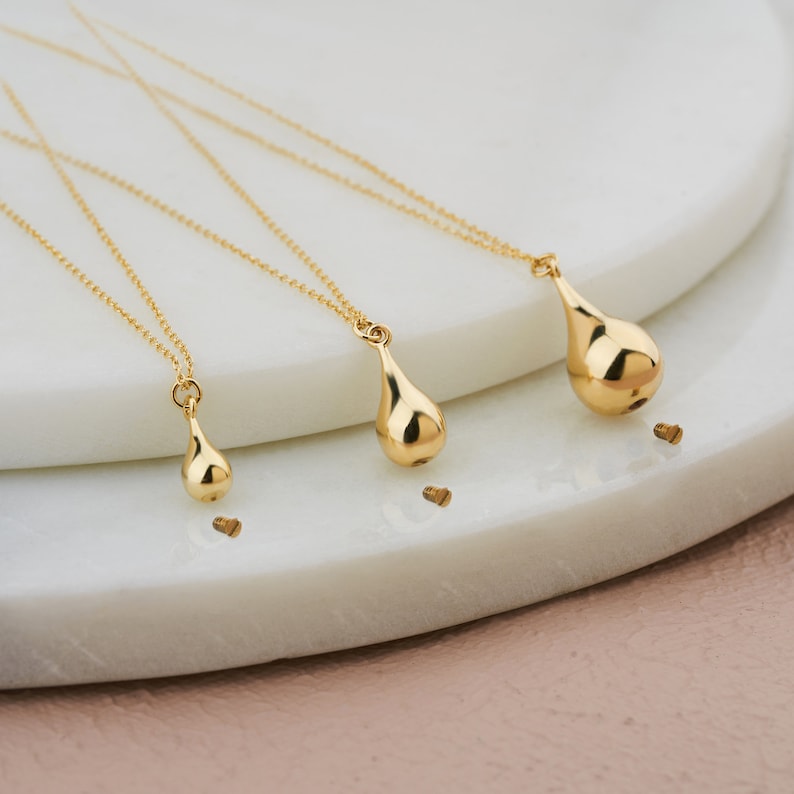 Three gold teardrop cremation urn necklaces in medium, large and xlarge sizes displayed on a white ceramic tray