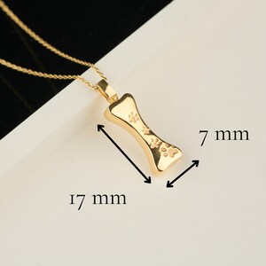 May include: A gold-colored pendant necklace featuring a bone-shaped charm with paw print details. The charm measures 17 mm long and 7 mm wide. The necklace has a delicate gold chain.