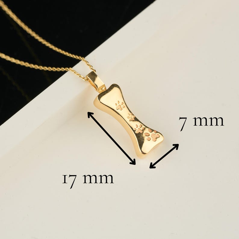 May include: A gold-colored pendant necklace featuring a bone-shaped charm with paw print details. The charm measures 17 mm long and 7 mm wide. The necklace has a delicate gold chain.