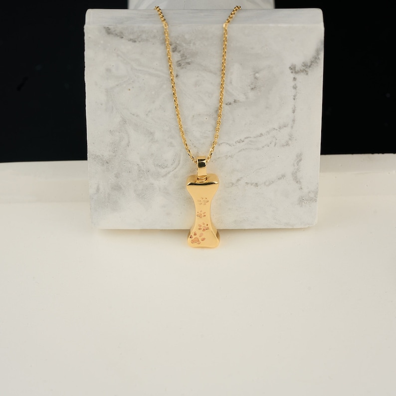 May include: A gold-colored bone-shaped pendant necklace with a delicate chain. The pendant features three paw prints. The necklace is displayed on a white and gray surface, with a black background.