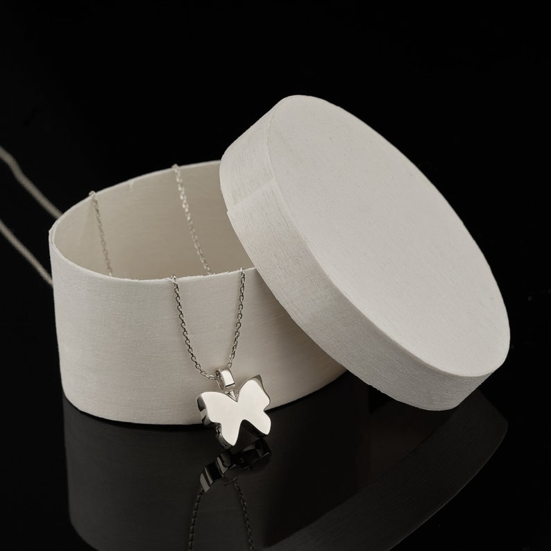 Puede incluir: Un collar de plata con un colgante de mariposa en una caja redonda blanca con la tapa abierta. El collar est&aacute; hecho de una cadena de plata delicada con un peque&ntilde;o dije de mariposa.