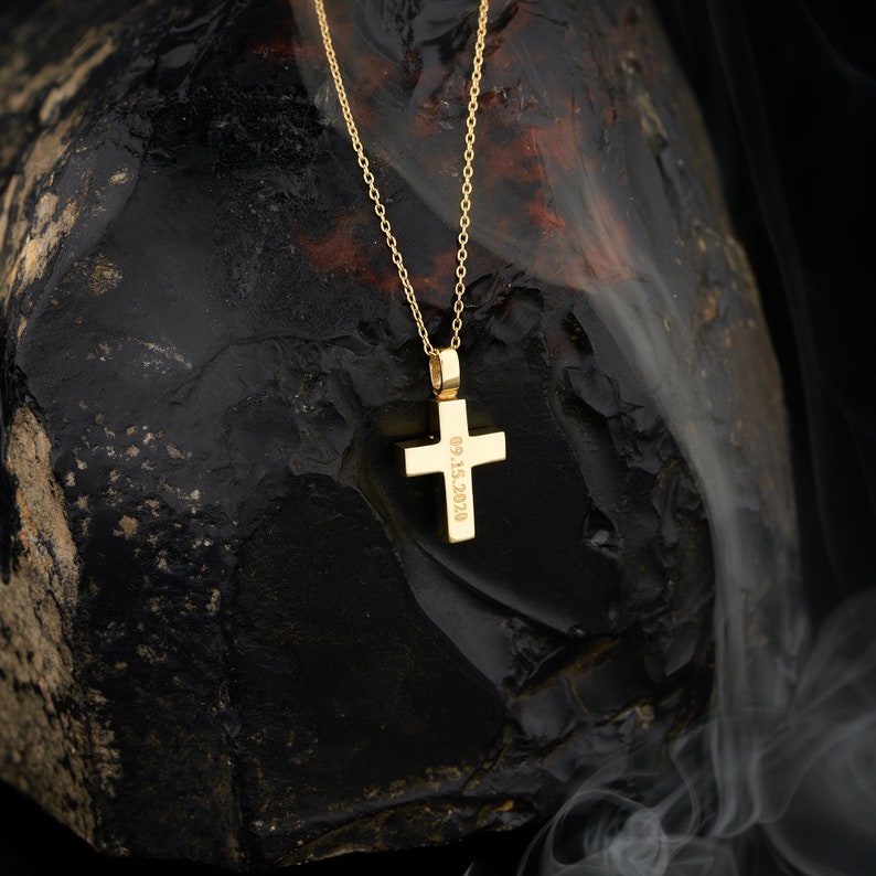 May include: A gold cross necklace with a date engraved on the back. The necklace is resting on a dark gray rock.