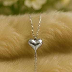 May include: A silver heart pendant necklace. The heart-shaped pendant is smooth and reflective, suspended from a delicate chain. The necklace hangs against a soft, yellow background, with blurred white flowers in the background.