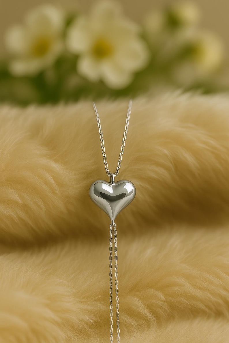 May include: A silver heart pendant necklace. The heart-shaped pendant is smooth and reflective, suspended from a delicate chain. The necklace hangs against a soft, yellow background, with blurred white flowers in the background.