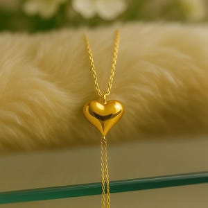 May include: A gold-colored heart pendant necklace. The heart-shaped pendant hangs from a delicate chain and features a smooth, polished surface. The necklace is displayed on a soft, cream-colored surface, with blurred white flowers in the background.