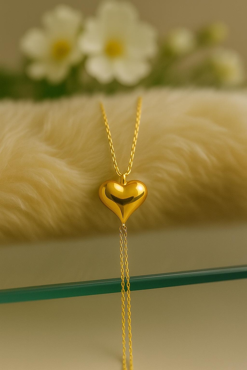 May include: A gold-colored heart pendant necklace. The heart-shaped pendant hangs from a delicate chain and features a smooth, polished surface. The necklace is displayed on a soft, cream-colored surface, with blurred white flowers in the background.