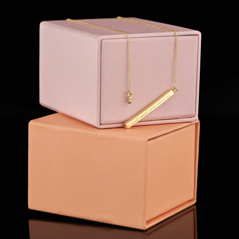 May include: A gold bar necklace with a delicate chain and the inscription "Still need you close to" on a pink and peach box.