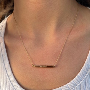 May include: A gold chain necklace with a thin bar pendant that says "My angel" on it.