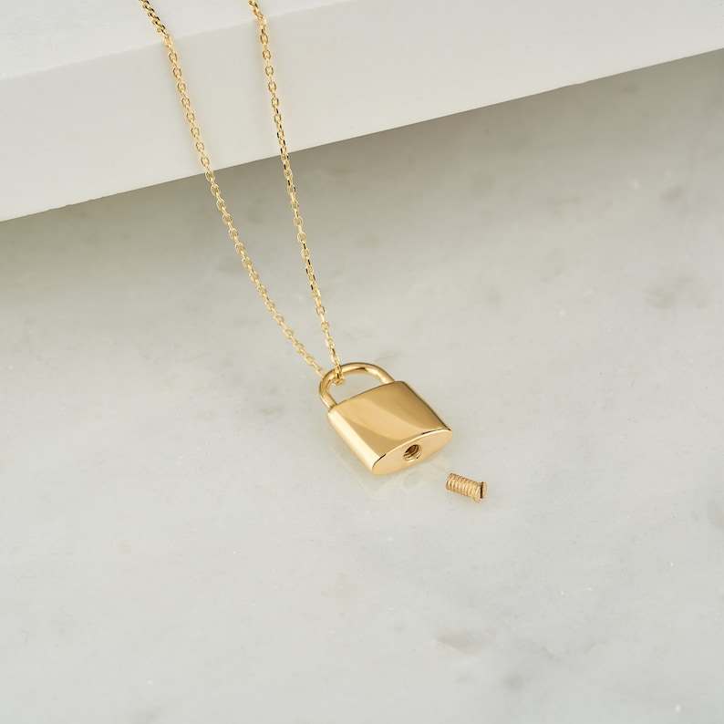 May include: A gold chain necklace with a small gold padlock pendant. The padlock is open and has a small screw on the side.