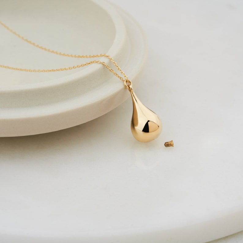 Gold cremation teardrop pendant resting on white ceramic tray with delicate gold chain
