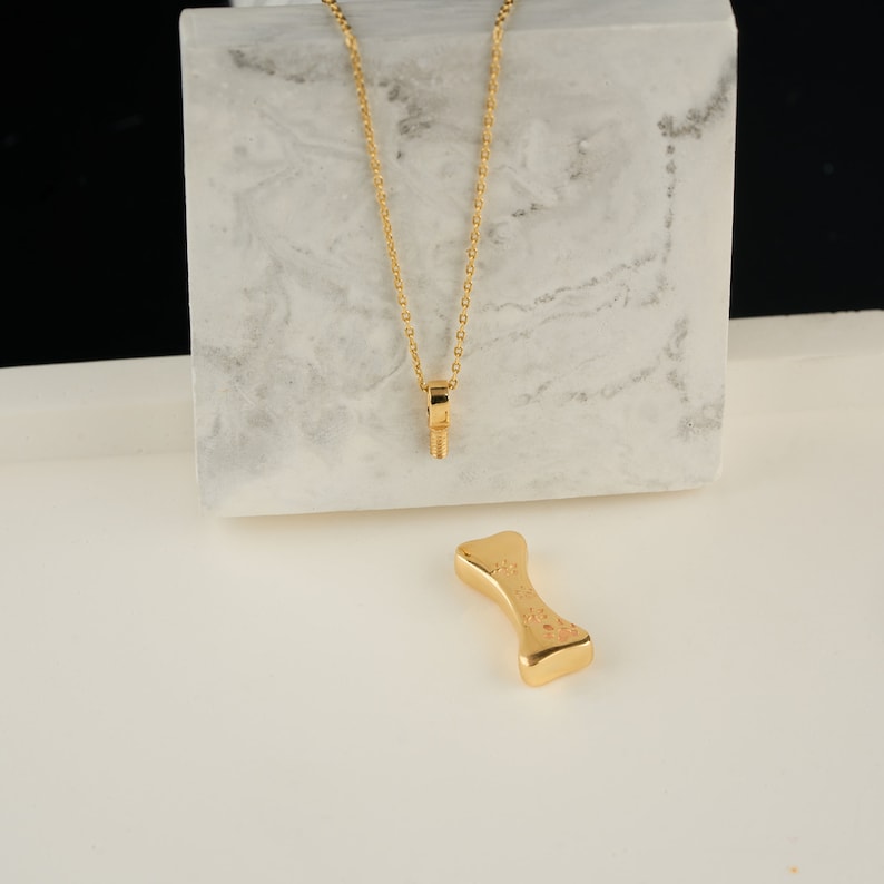 May include: A gold-colored necklace with a pendant shaped like a bolt. A gold-colored dog bone-shaped charm is also present. The items are displayed on a white surface with a marble-patterned block in the background.