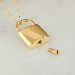 May include: A gold padlock necklace with a screw-on lid. The necklace is open and the lid is off, revealing the inside of the padlock.
