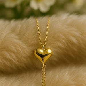 May include: A gold-colored heart pendant necklace. The heart-shaped pendant is smooth and reflective, suspended from a delicate chain. The necklace hangs against a soft, textured background, with blurred white flowers in the background.