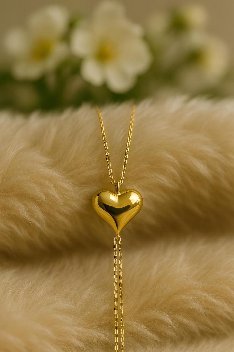 May include: A gold-colored heart pendant necklace. The heart-shaped pendant is smooth and reflective, suspended from a delicate chain. The necklace hangs against a soft, textured background, with blurred white flowers in the background.