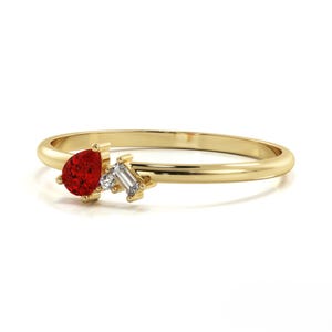 May include: A delicate gold ring featuring a pear-shaped red gemstone and two smaller, rectangular white gemstones. The ring has a thin band and a cluster design, perfect for everyday wear or special occasions.
