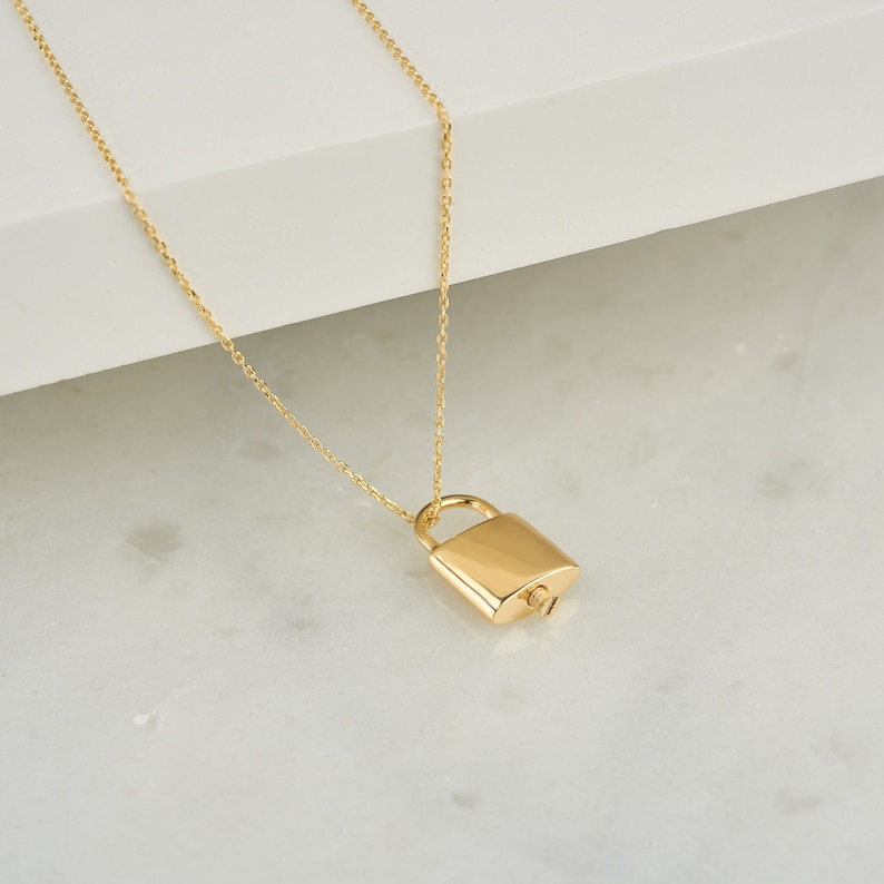 May include: A gold chain necklace with a padlock pendant. The padlock is a simple design with a rounded shape.