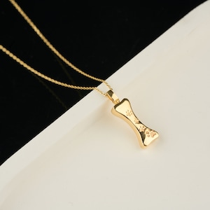 May include: A gold-colored necklace with a bone-shaped pendant. The pendant features paw print designs and hangs from a delicate gold chain. The necklace is displayed on a white surface with a black background.