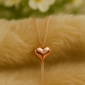 May include: A rose gold heart pendant necklace. The heart-shaped pendant is smooth and reflective, suspended from a delicate chain. The necklace hangs against a soft, yellow background, with blurred white flowers in the background.