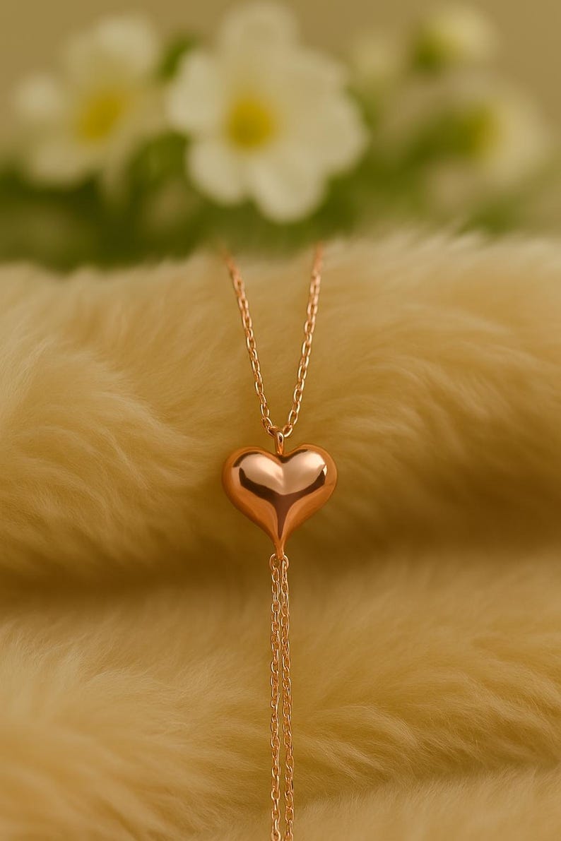 May include: A rose gold heart pendant necklace. The heart-shaped pendant is smooth and reflective, suspended from a delicate chain. The necklace hangs against a soft, yellow background, with blurred white flowers in the background.