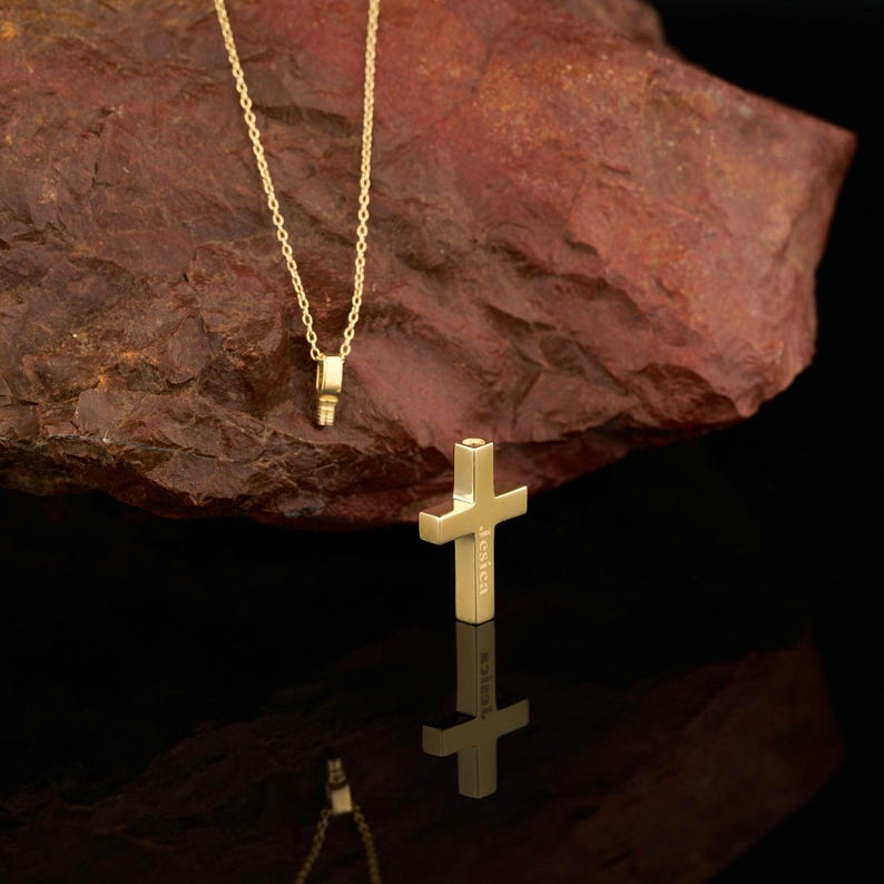 May include: A gold chain necklace with a small, rectangular pendant. The pendant is a cross with the word "Jessica" engraved on it.