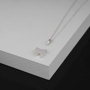 Puede incluir: Un collar con un colgante de mariposa de plata sobre una superficie blanca. El colgante es un dise&ntilde;o simple y elegante con una cadena delicada.