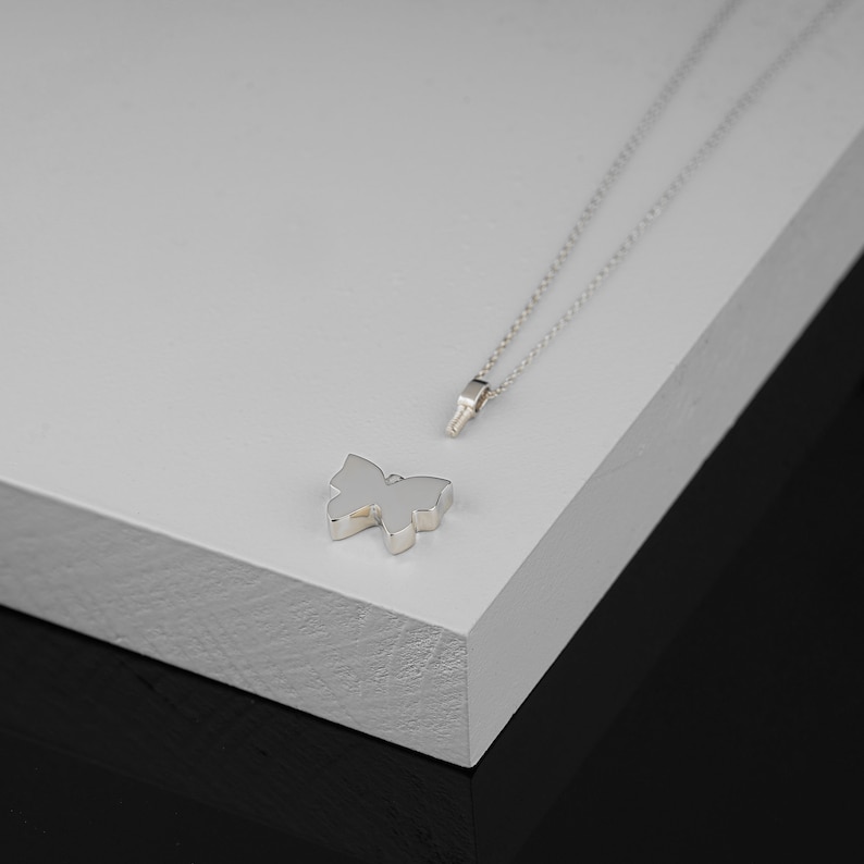 Puede incluir: Un collar con un colgante de mariposa de plata sobre una superficie blanca. El colgante es un dise&ntilde;o simple y elegante con una cadena delicada.