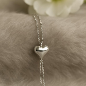 May include: A silver heart pendant necklace. The heart-shaped pendant hangs from a delicate chain, with two additional chains extending below the heart. The necklace is displayed on a soft, textured surface, with a blurred white flower in the background.