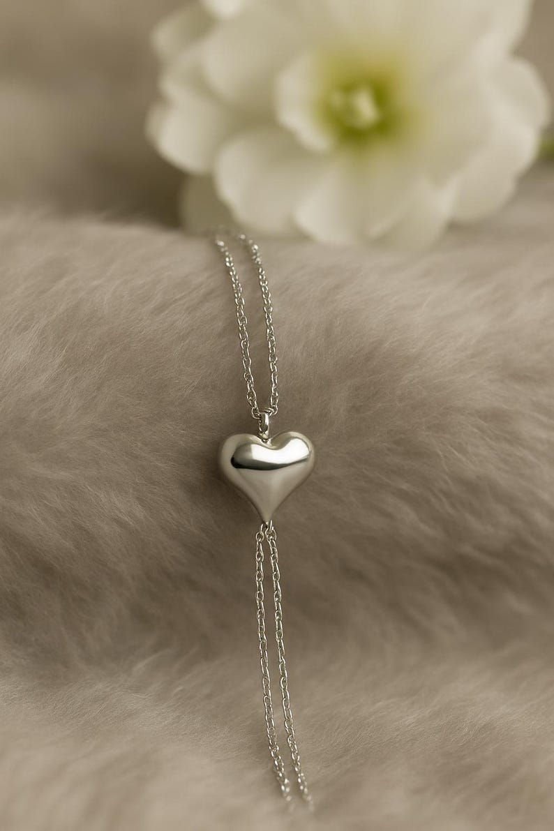 May include: A silver heart pendant necklace. The heart-shaped pendant hangs from a delicate chain, with two additional chains extending below the heart. The necklace is displayed on a soft, textured surface, with a blurred white flower in the background.