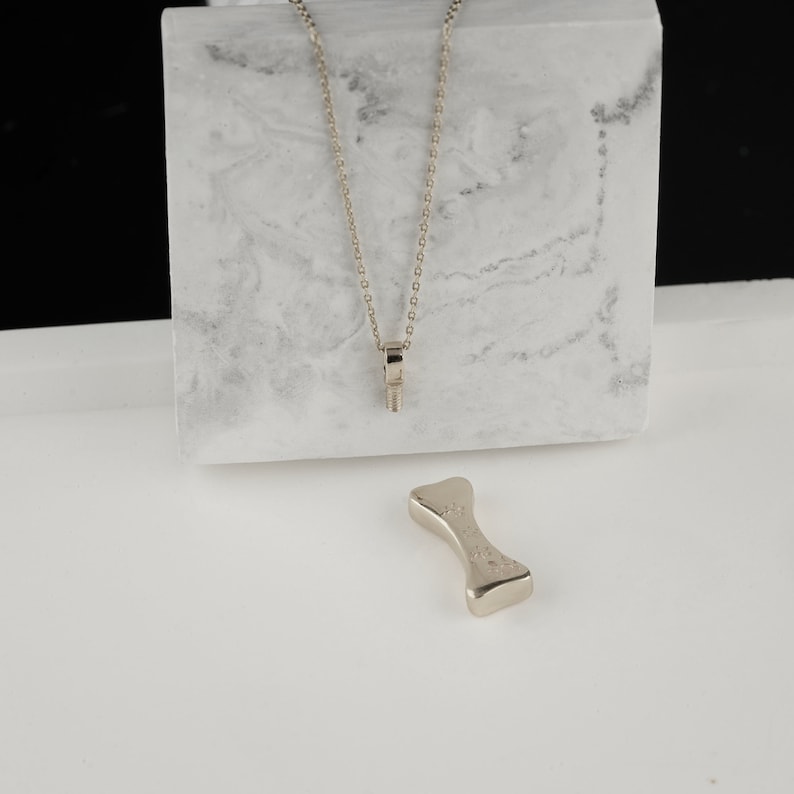May include: A silver-toned necklace with a bolt pendant hangs in front of a marble block. A matching bone-shaped object rests below. The jewelry is displayed on a white surface with a black background.
