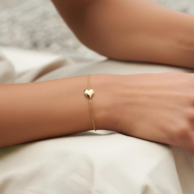 May include: A delicate gold bracelet featuring a heart-shaped charm. The bracelet is a thin chain with a polished heart pendant. The jewelry is displayed on a wrist, showcasing the bracelet's design and color.