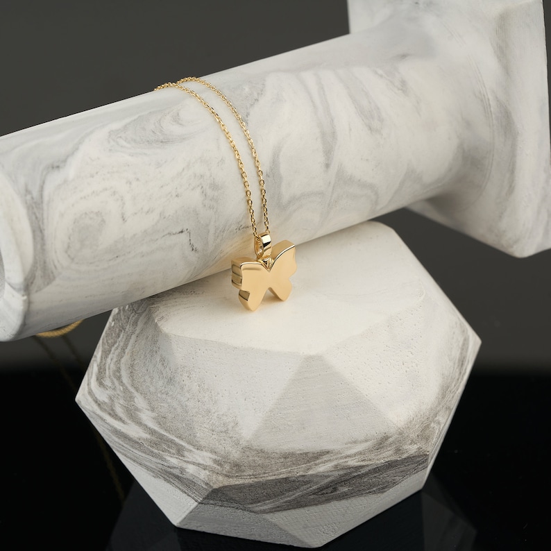 Puede incluir: Un collar con un colgante de mariposa de oro sobre una superficie de m&aacute;rmol blanco. El colgante es un dise&ntilde;o simple y elegante con una cadena delicada.