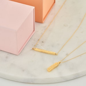 May include: Two gold necklaces on a white marble surface. One necklace has a bar pendant with the inscription "I will wear you close to my heart always". The other necklace has a rectangular pendant.