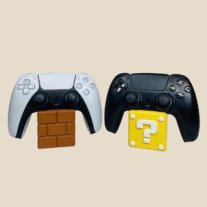 May include: Two video game controllers, one white and one black, are displayed on stands. The white controller is on a brown brick stand, and the black controller is on a yellow question mark stand.