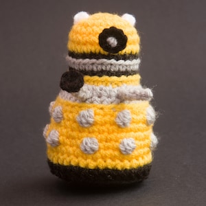 May include: A yellow crocheted toy with black and grey details. The toy is shaped like a robot with a round body and a dome-shaped head.