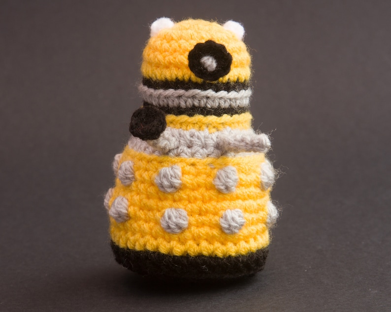 May include: A yellow crocheted toy with black and grey details. The toy is shaped like a robot with a round body and a dome-shaped head.