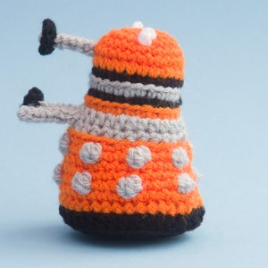 May include: A crocheted orange and grey toy with black accents and white dots. The toy is shaped like a robot with two arms and a dome-shaped head.
