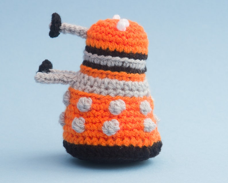 May include: A crocheted orange and grey toy with black accents and white dots. The toy is shaped like a robot with two arms and a dome-shaped head.