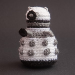 May include: A crocheted white and grey robot toy with black details. The robot has a round body with a dome-shaped head and a single eye.