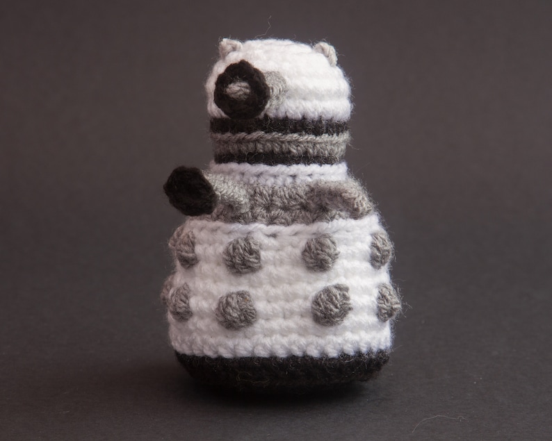 May include: A crocheted white and grey robot toy with black details. The robot has a round body with a dome-shaped head and a single eye.