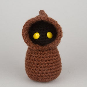 May include: A brown crocheted plush toy with a pointed hood and yellow eyes. The toy is shaped like a small person.