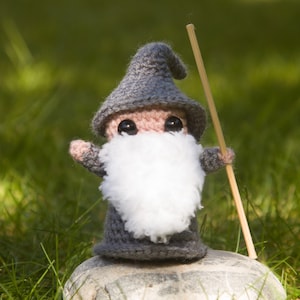 May include: A small gray crocheted gnome with a white beard and a wooden stick in its hand. The gnome is standing on a gray rock.