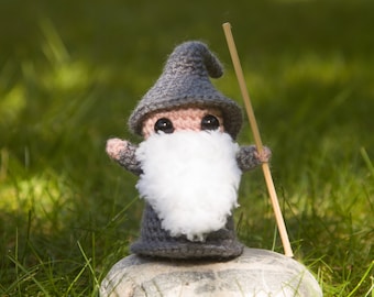 May include: A small gray crocheted gnome with a white beard and a wooden stick in its hand. The gnome is standing on a gray rock.