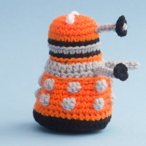 May include: A crocheted orange and grey toy with black accents and white dots. The toy is shaped like a robot with a round head and a long, thin body.