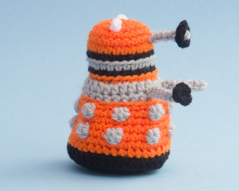 May include: A crocheted orange and grey toy with black accents and white dots. The toy is shaped like a robot with a round head and a long, thin body.