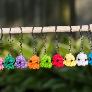 May include: Eight crochet octopus keyrings in a row, each a different colour: pink, green, purple, orange, green, red, yellow, white, and blue. They are suspended from a wooden dowel against a blurred green backdrop.