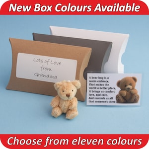 May include: A small teddy bear sits on a blue surface next to a brown, grey, and white pillow box. The box has a white label with the text "Lots of Love from Grandma". A clear plastic card with a teddy bear image and a poem about bear hugs is also visible.
