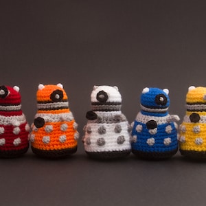 May include: Five crocheted toy robots in different colours, including red, orange, white, blue, and yellow. Each robot has a round body with black eyes and a black stripe around the middle.