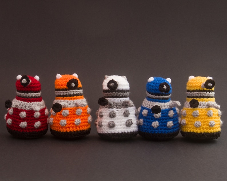 May include: Five crocheted toy robots in different colours, including red, orange, white, blue, and yellow. Each robot has a round body with black eyes and a black stripe around the middle.