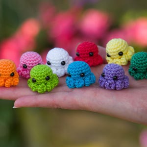 May include: Nine small, crocheted octopus toys in a variety of colours: orange, pink, white, red, yellow, green, blue, and purple. Each octopus has black eyes and eight tentacles. The toys are displayed on a hand, with a blurred background.